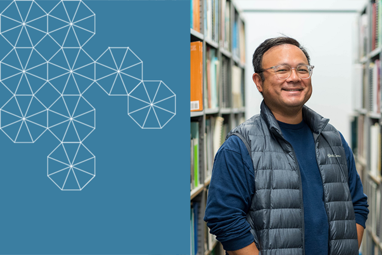 Mike Santos stands in a library with books to either side of him, smiling and wearing glasses