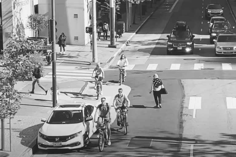 City street scene in black and white with people walking and biking, while two lanes of cars wait at an intersection 