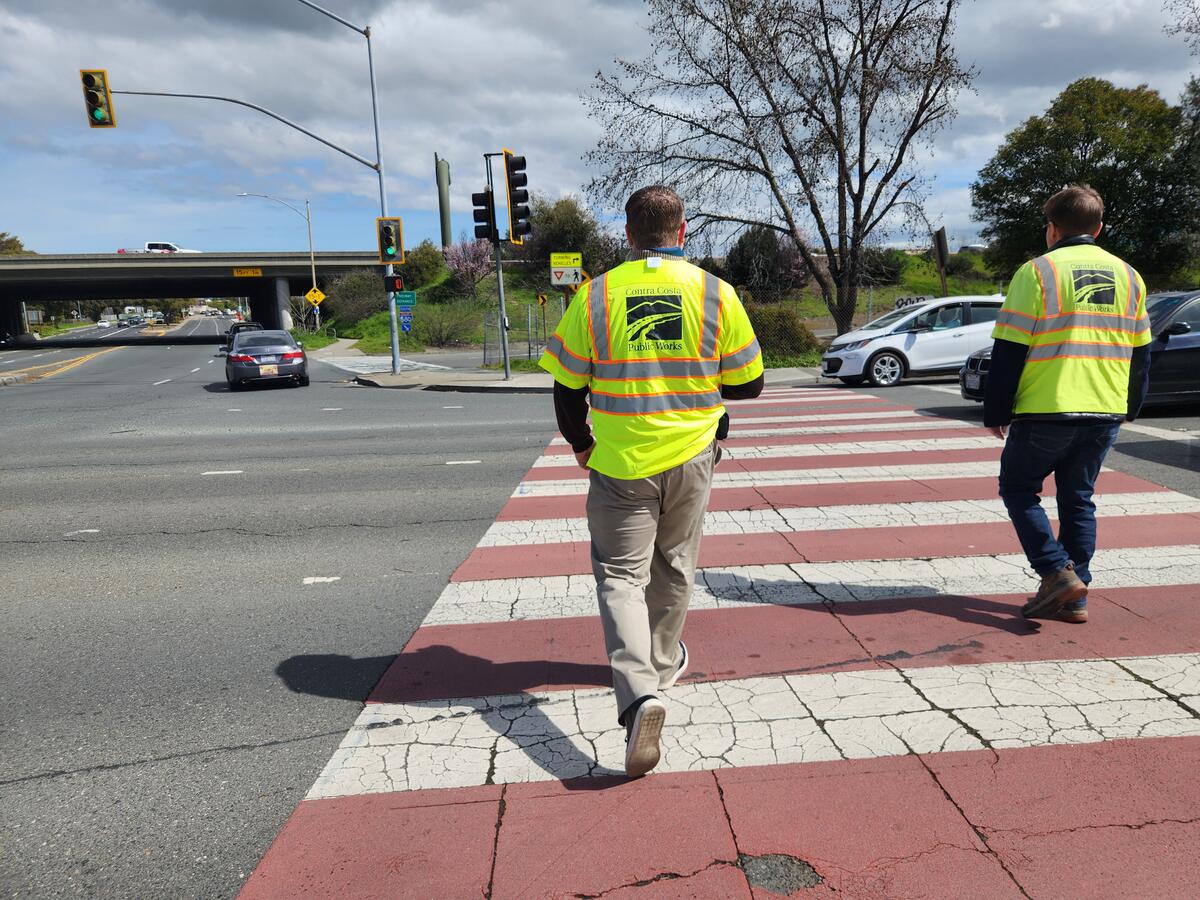 Two people wearing safety vests labelled "Contra Costa Public Works" are walking along a crosswalk