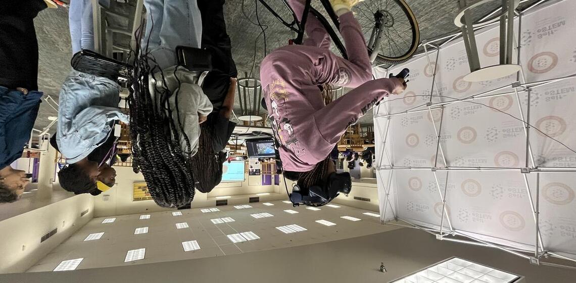 A girl is riding the bicycle simulator, while a group of girls and a woman watch