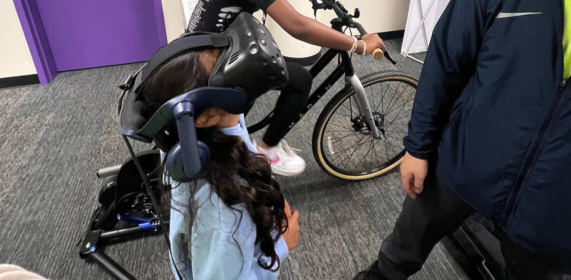 An older girl pedals on the bicycle simulator while a younger girl wears the virtual reality headse