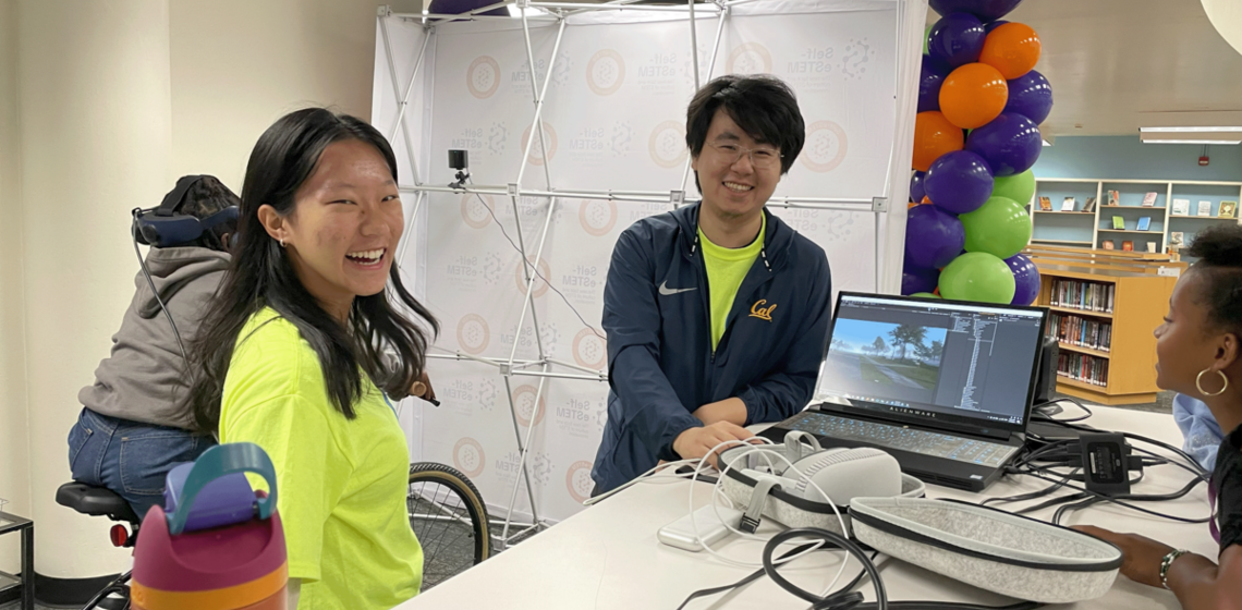 Florance and Han are sitting and smiling at the camera. In the background, one girl is pedaling on the bicycle simulator and ano