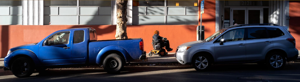 A person in a motorized wheelchair on a sidewalk in between a truck and SUVnd 